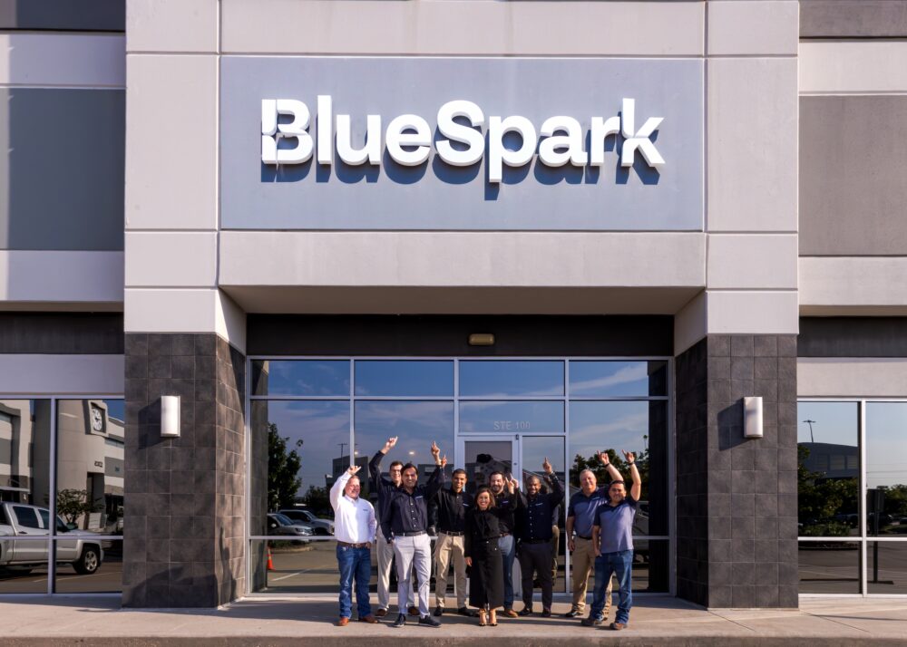BlueSpark CEO Kahina, CTO Matt and team point at the BlueSpark sign on the new Houston HQ