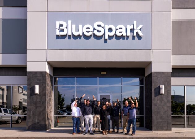 BlueSpark CEO Kahina, CTO Matt and team point at the BlueSpark sign on the new Houston HQ